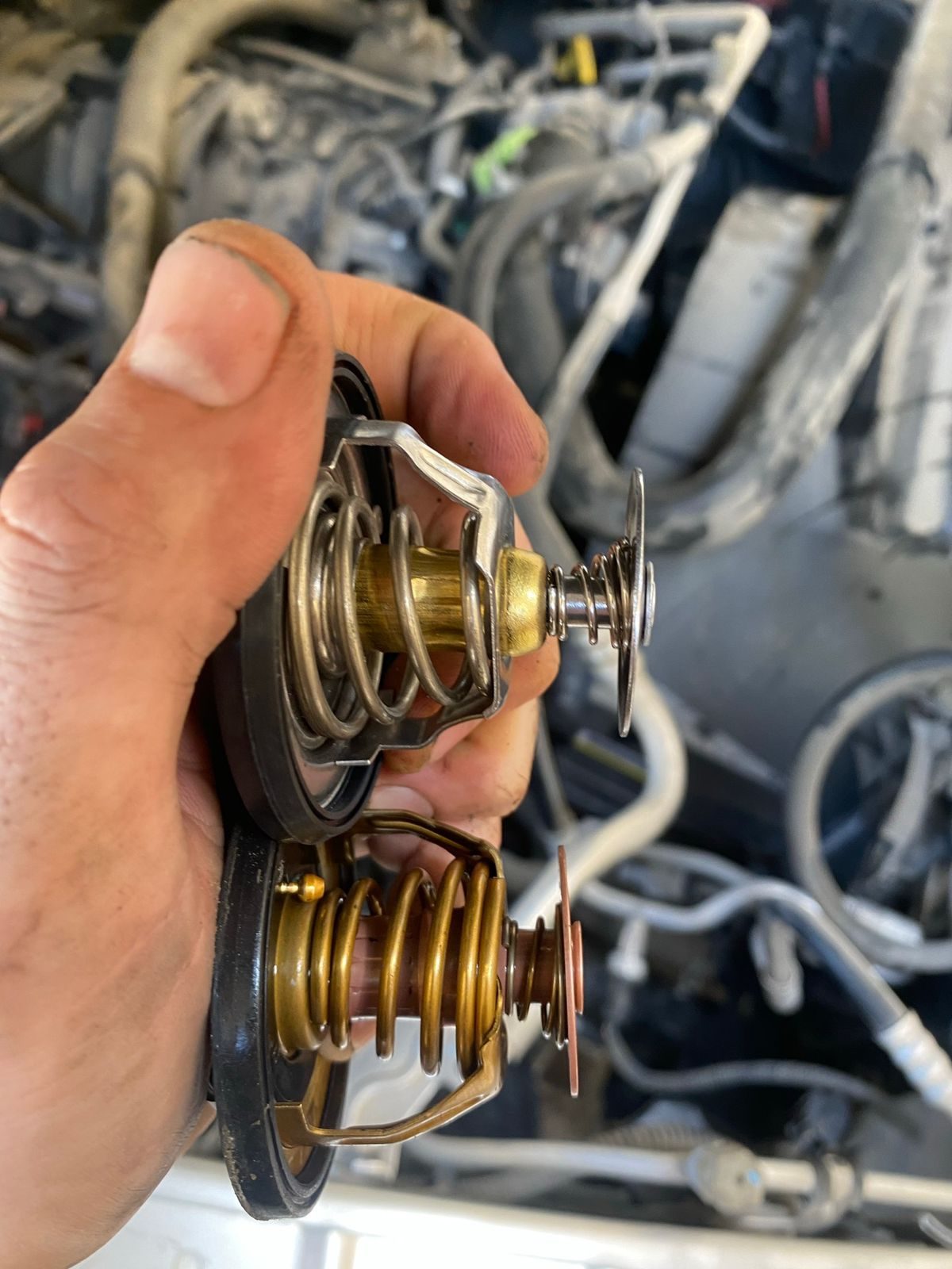 Two thermostats held side by side over the engine bay. Old vs replacement.