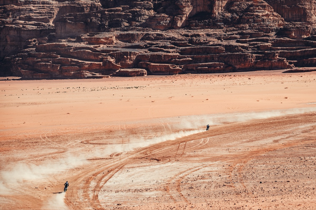 Wide pan of Wadi Rum cliff faces with two motorcycles tracing the desert floor in the distance.